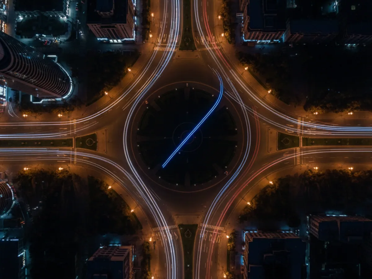A roundabout at night from above: light trails from cars and a blue beacon trail through the middle